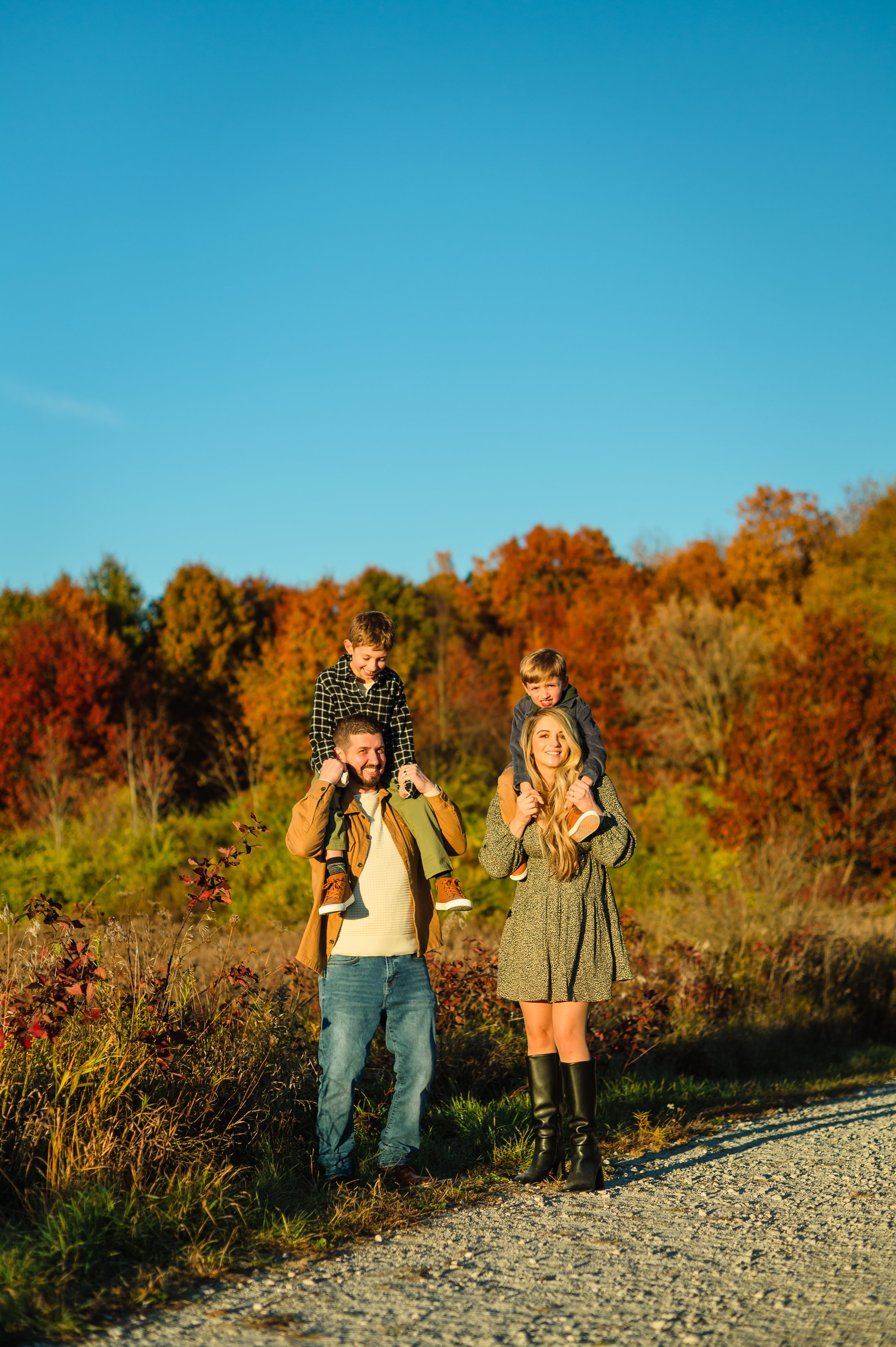 A smiling family of four enjoying an autumn walk in a park with colorful fall foliage, two children sitting on their parents' shoulders, one boy on his father’s shoulders and the same girl on his mother’s, all on a sunny day.