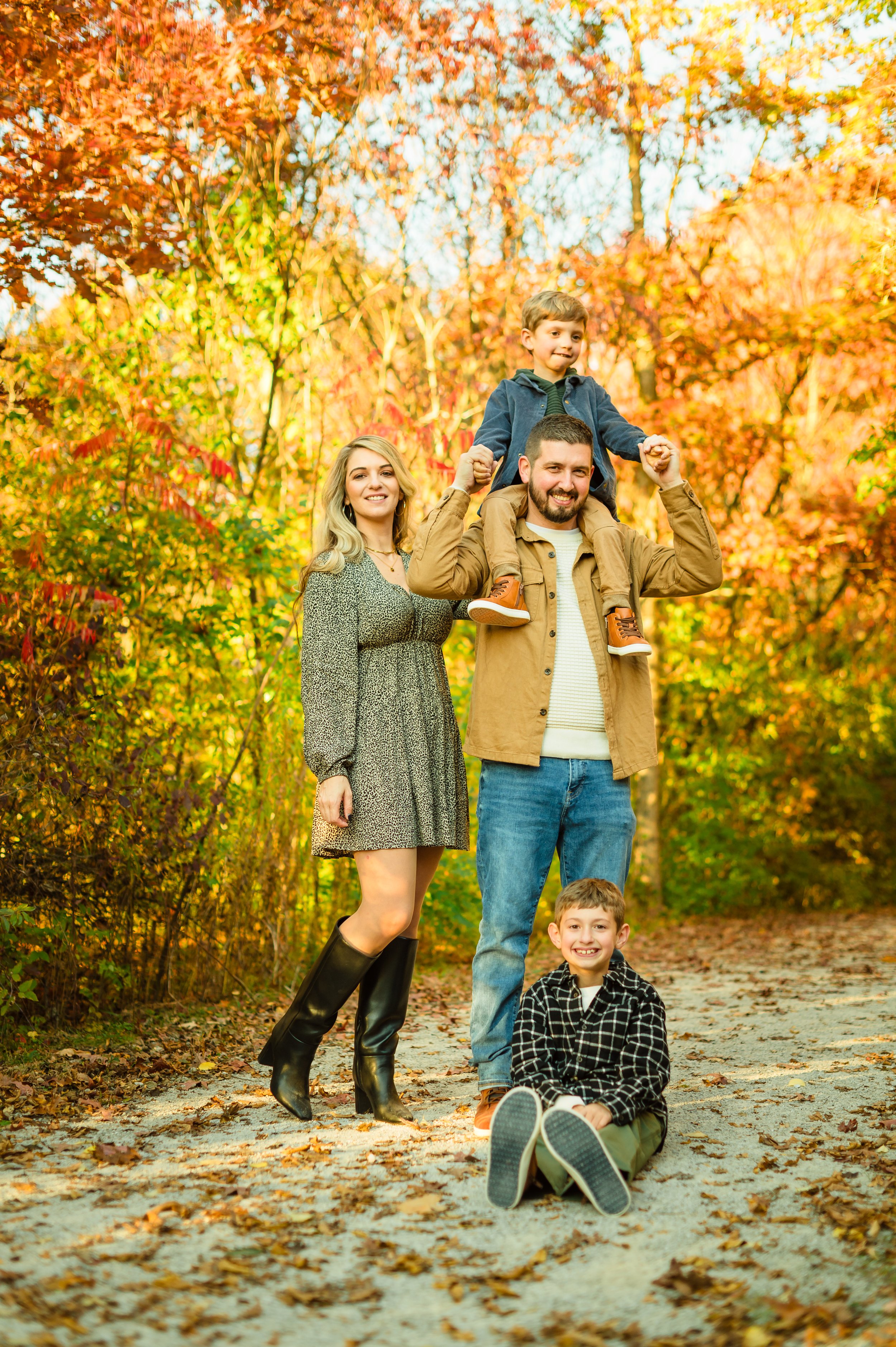 A family of four enjoying a fall day outdoors in a forest with colorful autumn leaves. The father is carrying a young boy on his shoulders, and the mother and older boy are standing beside them, smiling. The scene is vibrant with fall colors.