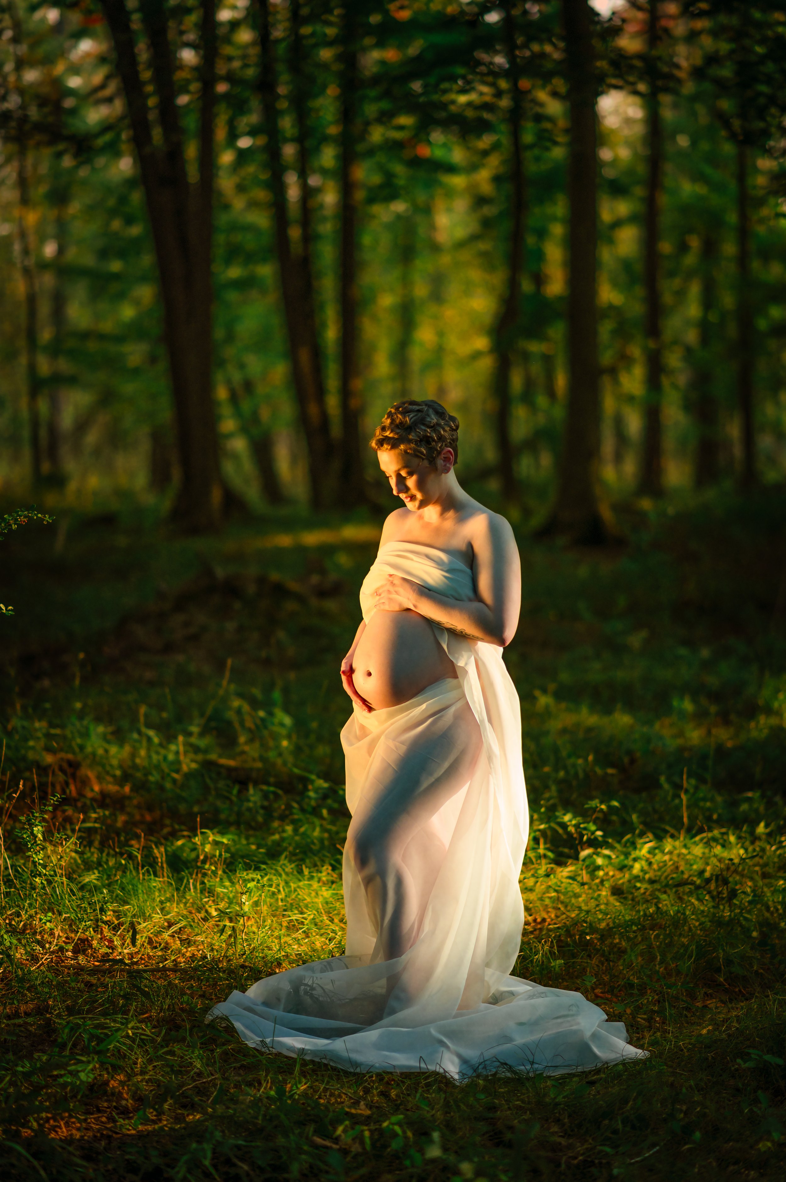 A pregnant woman in a flowing white dress standing outdoors in a forest at sunset, gently holding her belly and looking down.
