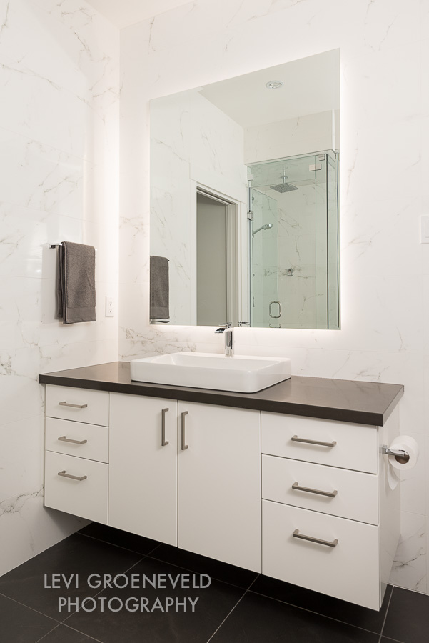 The modern master bathroom has clean lines with a floating countertop and backlit mirror. It features a beautiful grey quartz countertop and a square charcoal floor tile. 