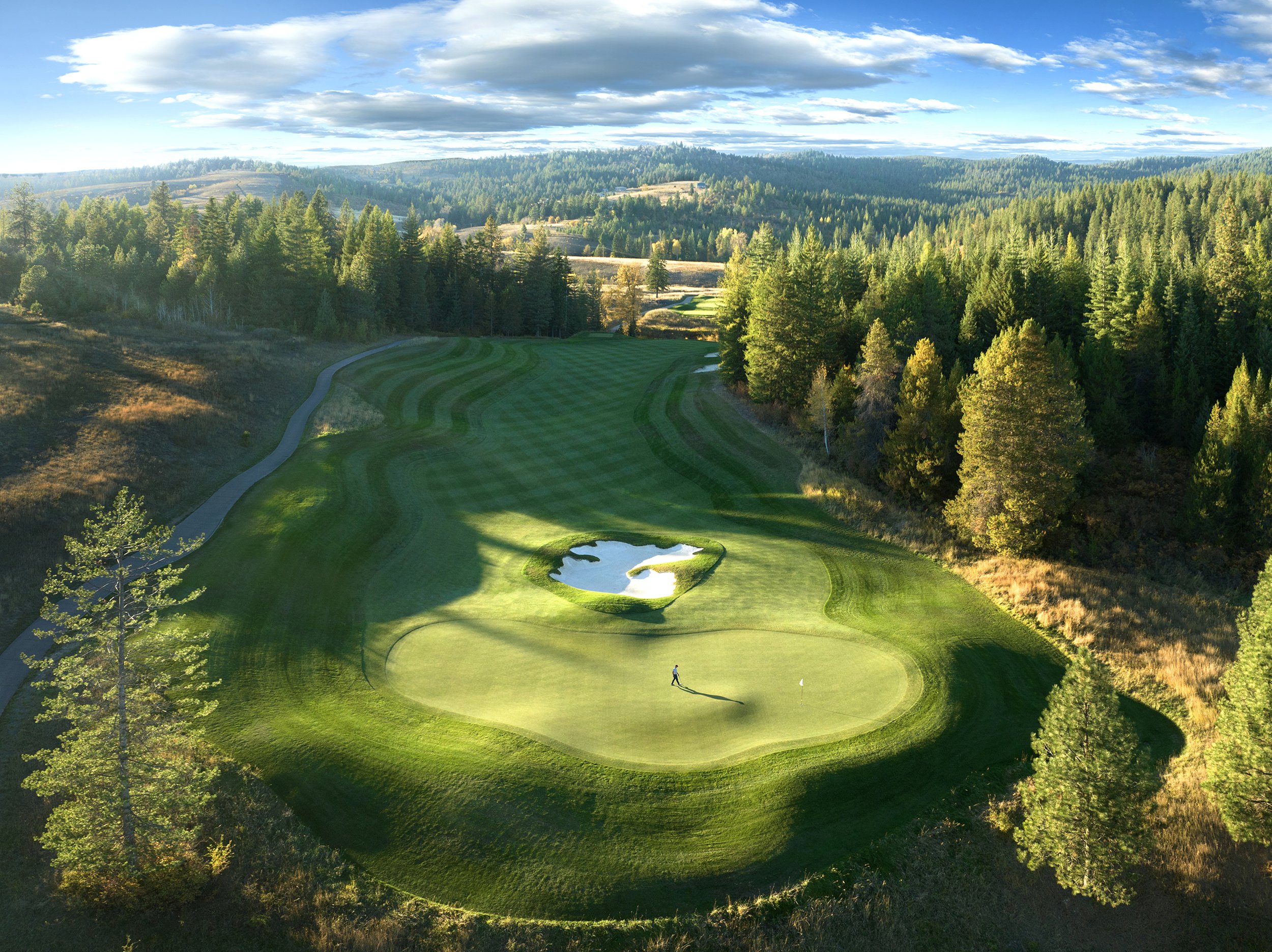 Aerial drone photograph of CDA National Golf Course showing a golfer walking on the putting green, with surrounding fairway, bunkers, and natural landscape visible from above