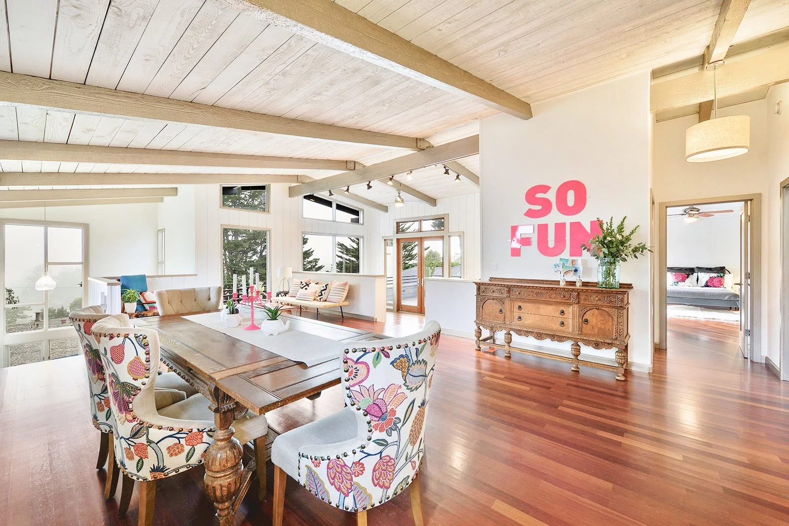 Bright, airy dining and living space with a wooden dining table, floral upholstered chairs, and a built-in bench with pillows. Large windows and a door lead outside. A vintage wooden sideboard with a vase of flowers and a wall with pink lettering that says 'SO FUN'. In the adjacent room, a glimpse of a bed with floral pillows is visible.