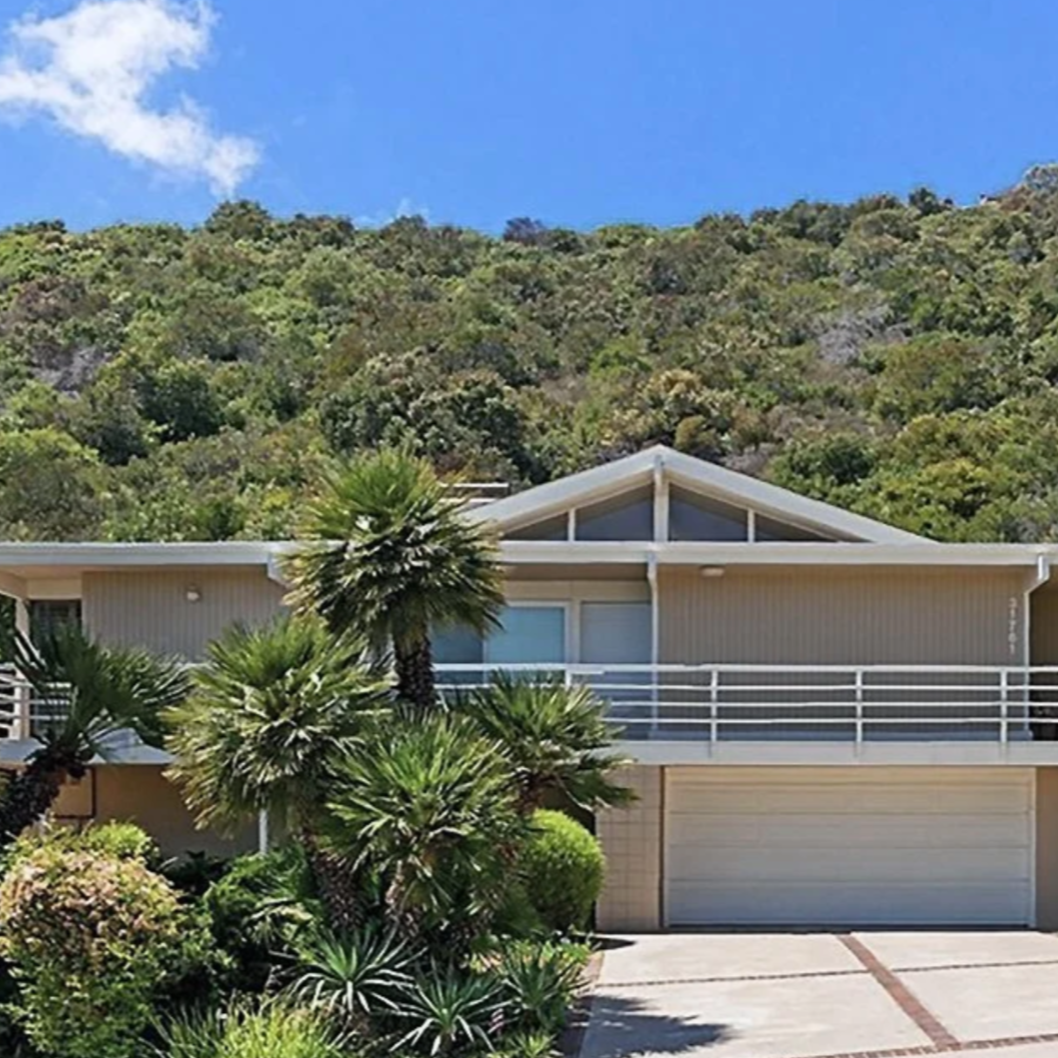 A mid-century modern house, surrounded by lush greenery and palm trees, with a green hillside and blue sky in the background.