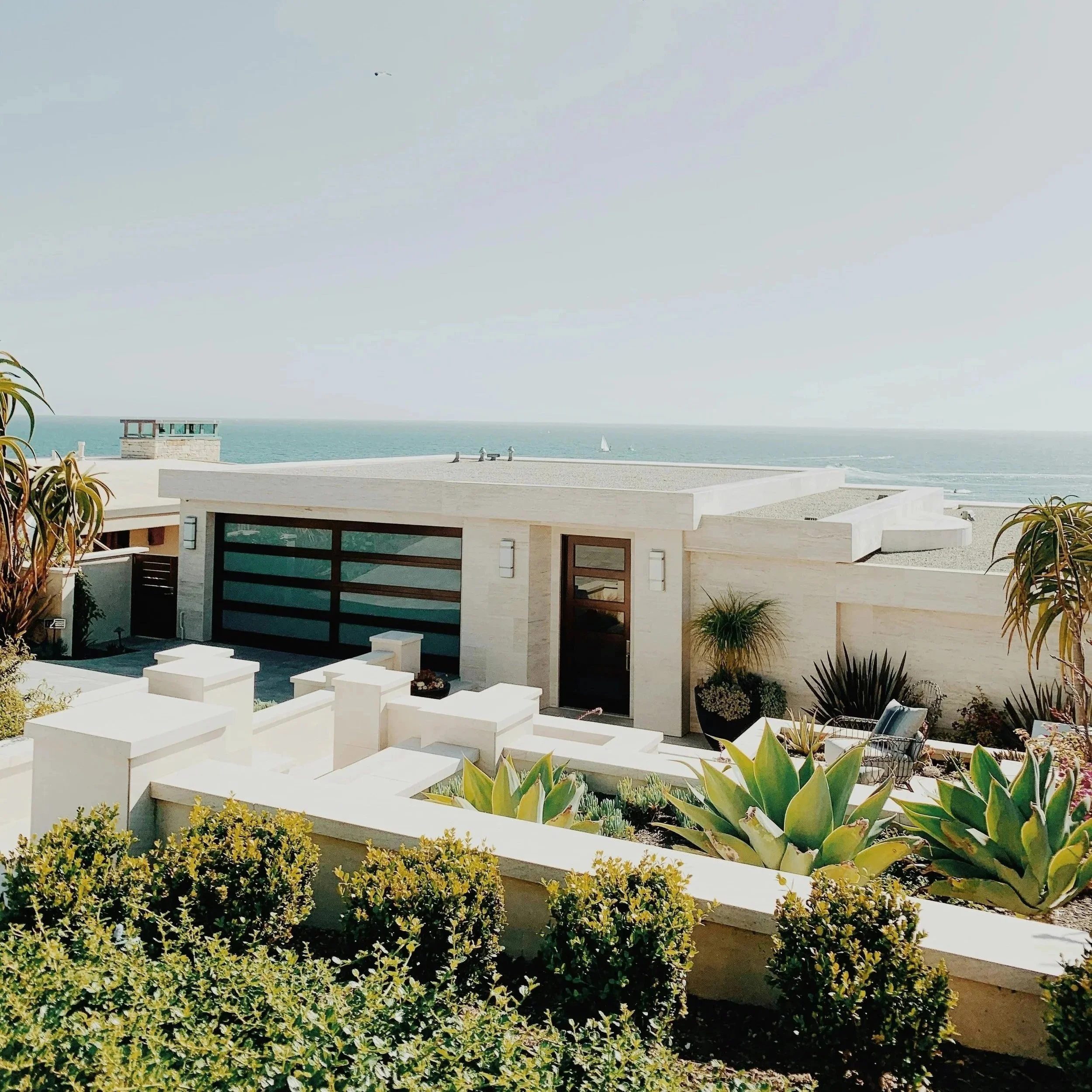 Modern house with a flat roof, large windows, and white exterior, overlooking the ocean with blue skies and a sailboat in the distance. There are plants and shrubs in the foreground.