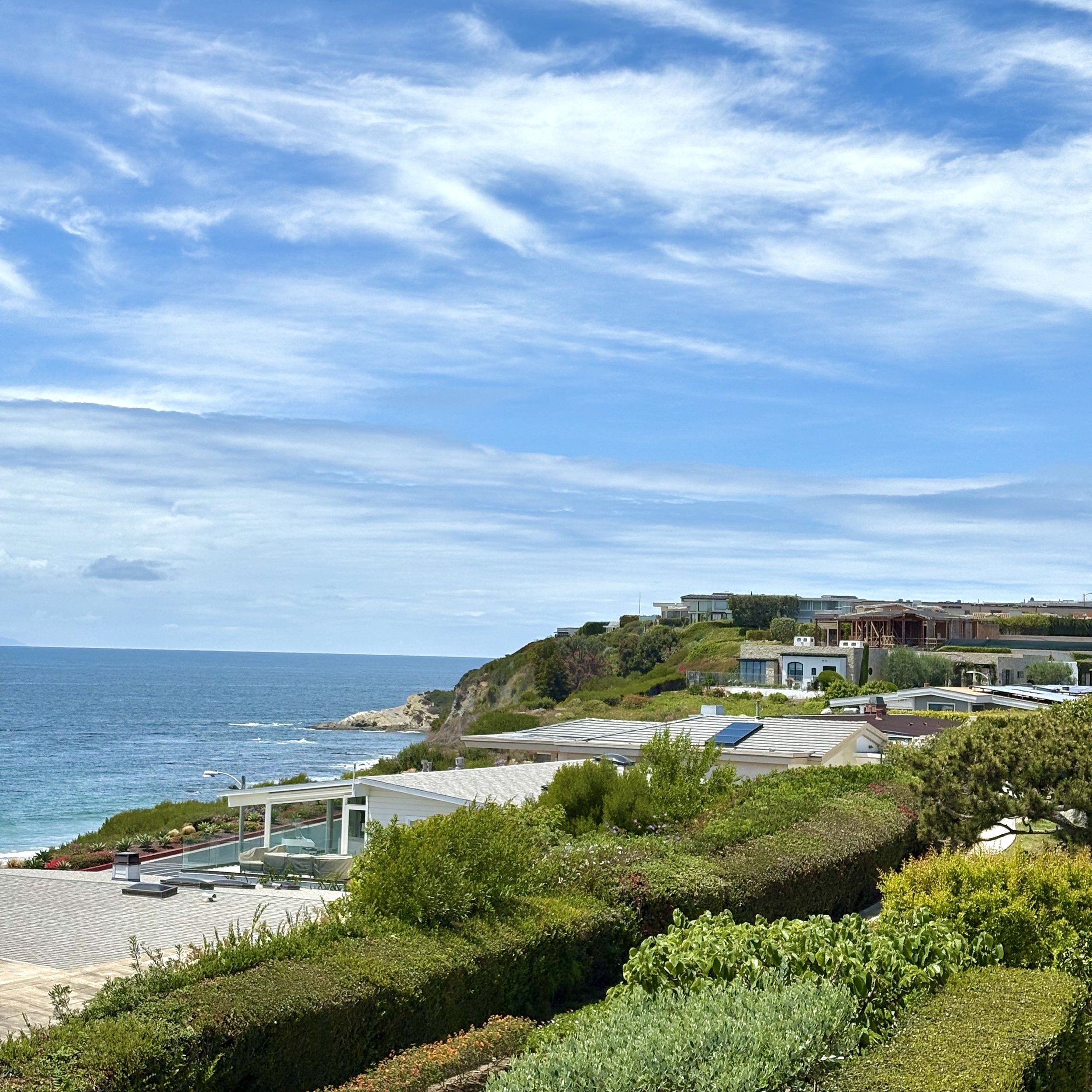 Scenic view of a coastal neighborhood with modern houses on a hill, lush greenery, and the ocean in the background under a blue sky with wispy clouds.