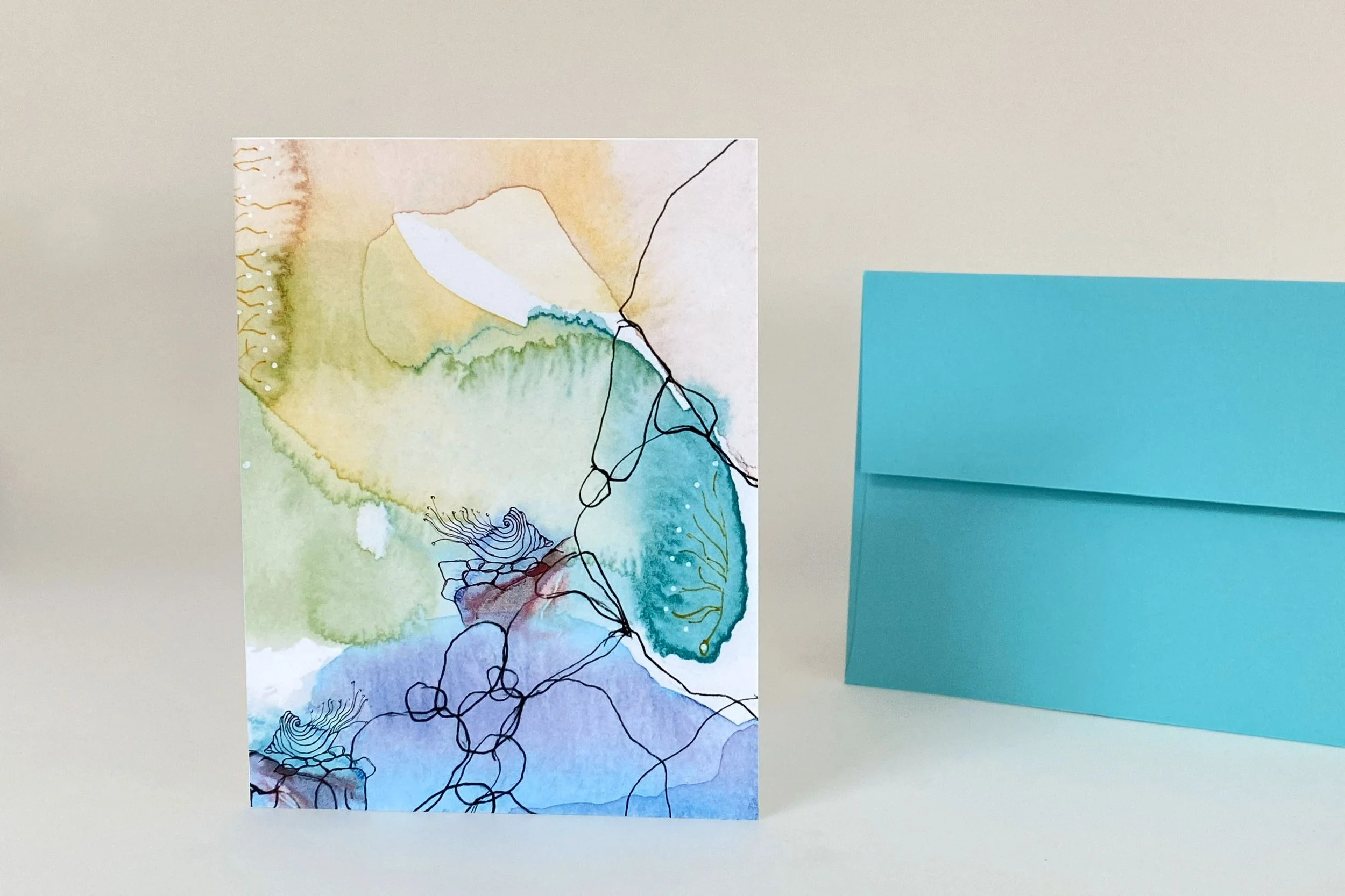 Abstract watercolor greeting card featuring layered blue, green, and earth-tone artwork with expressive line drawing, shown with teal envelope