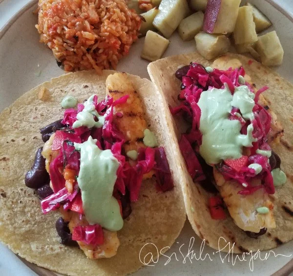 Image of a plate of 2 tacos with corn tortillas, pickled red cabbage, kidney beans, and grilled tempeh topped with creamy cilantro lime dressing. Also pictured on the plate is jollof rice and Japanese sweet potatoes. 