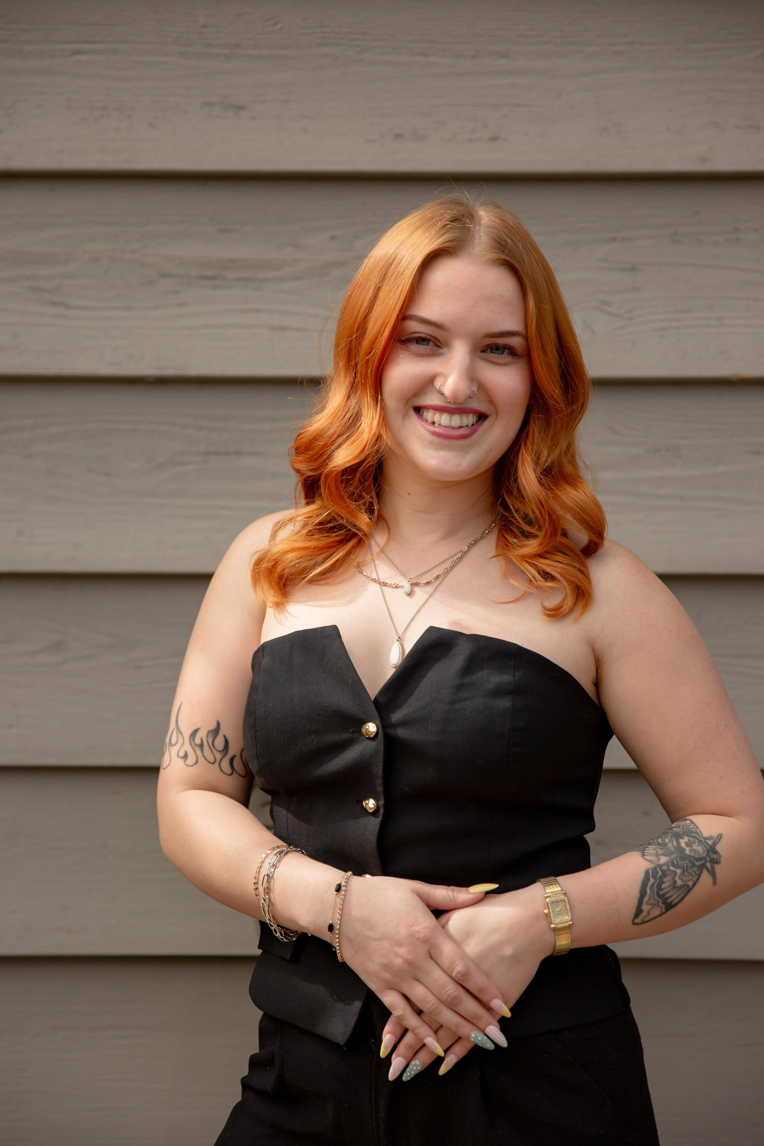 A woman with red hair, tattoos on her arms, and wearing a black strapless top stands in front of a gray wood-paneled wall. She is smiling and wearing jewelry, including necklaces, bracelets, and a gold watch.