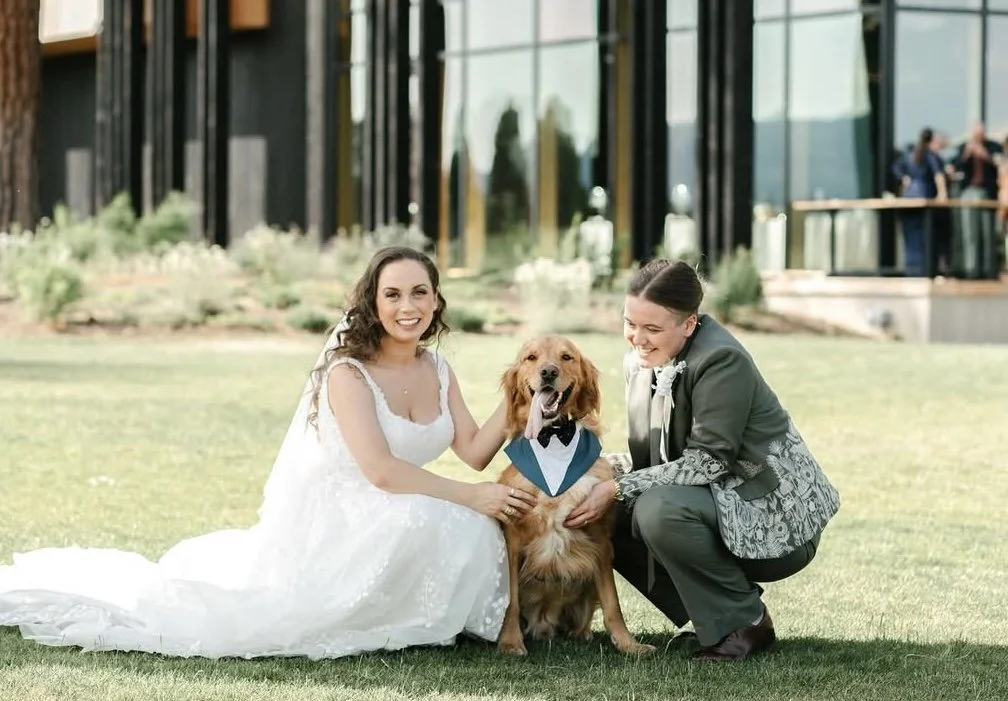 Two women and a golden retriever at a wedding outdoor ceremony, with modern building in background.
