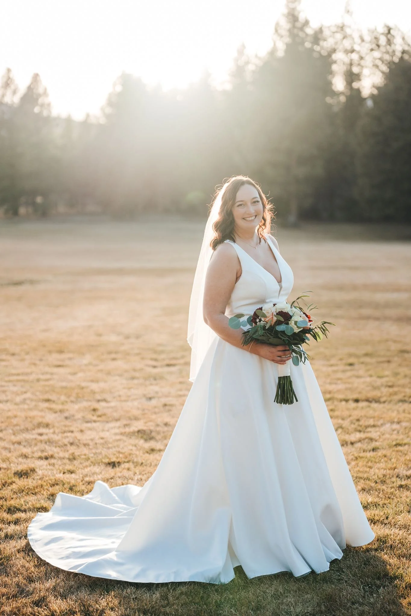 A smiling woman in a white wedding dress holding a bouquet of flowers standing outdoors on a grassy field in the light of the setting sun, with trees blurred in the background.