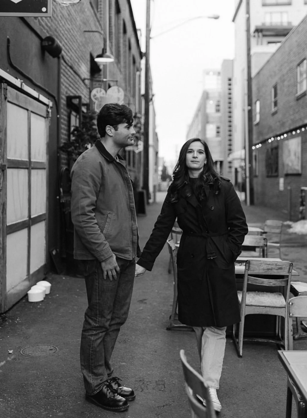 A black and white photo of a man and woman standing on a city sidewalk, holding hands. The man is on the left, wearing a jacket and jeans, and looking at the woman. The woman is on the right, wearing a long coat, with her other hand in her pocket, lo