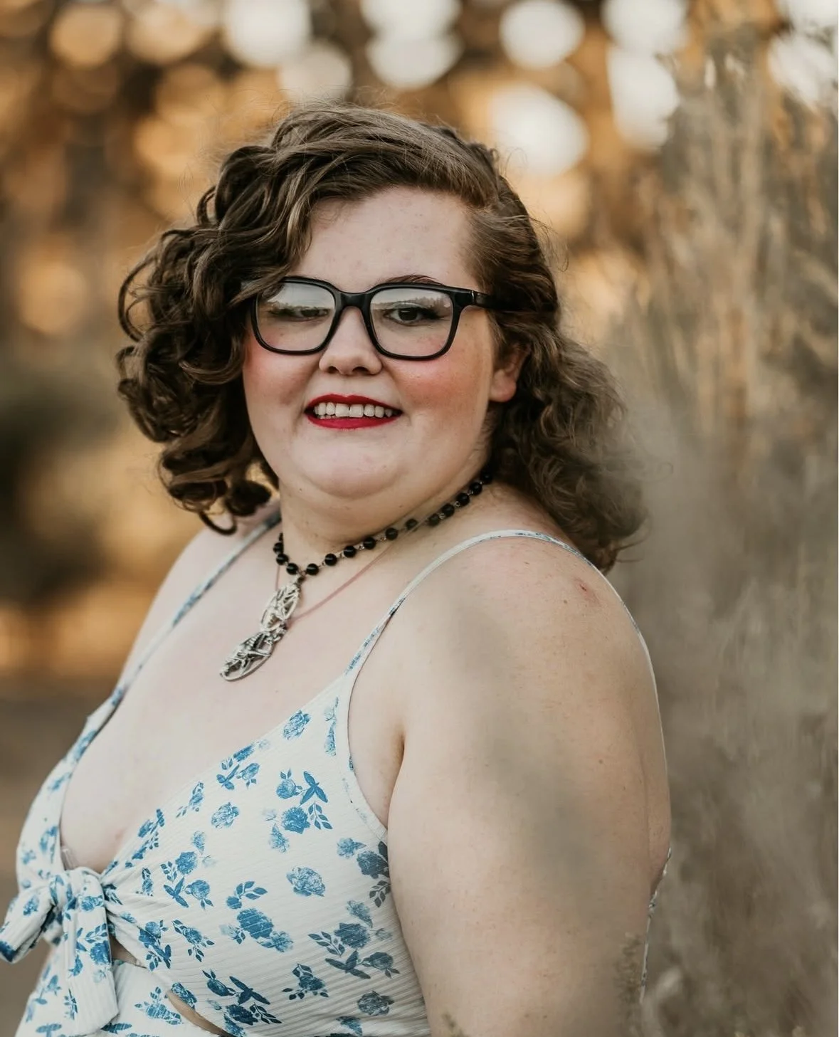 A woman with curly brown hair, glasses, red lipstick, and a floral dress, smiling outdoors with blurred trees in the background.