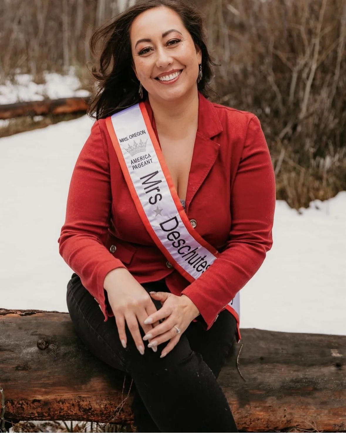 A woman sitting outdoors on a log in a snowy landscape, wearing a red jacket and a sash that reads 'Mrs. Denton' and 'Mrs. Oregon.' She is smiling at the camera.