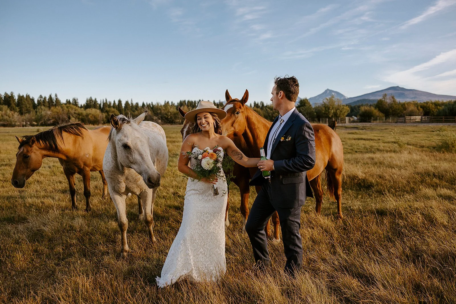 A bride and groom sharing a moment with horses in a scenic field during sunset, with mountains in the background.