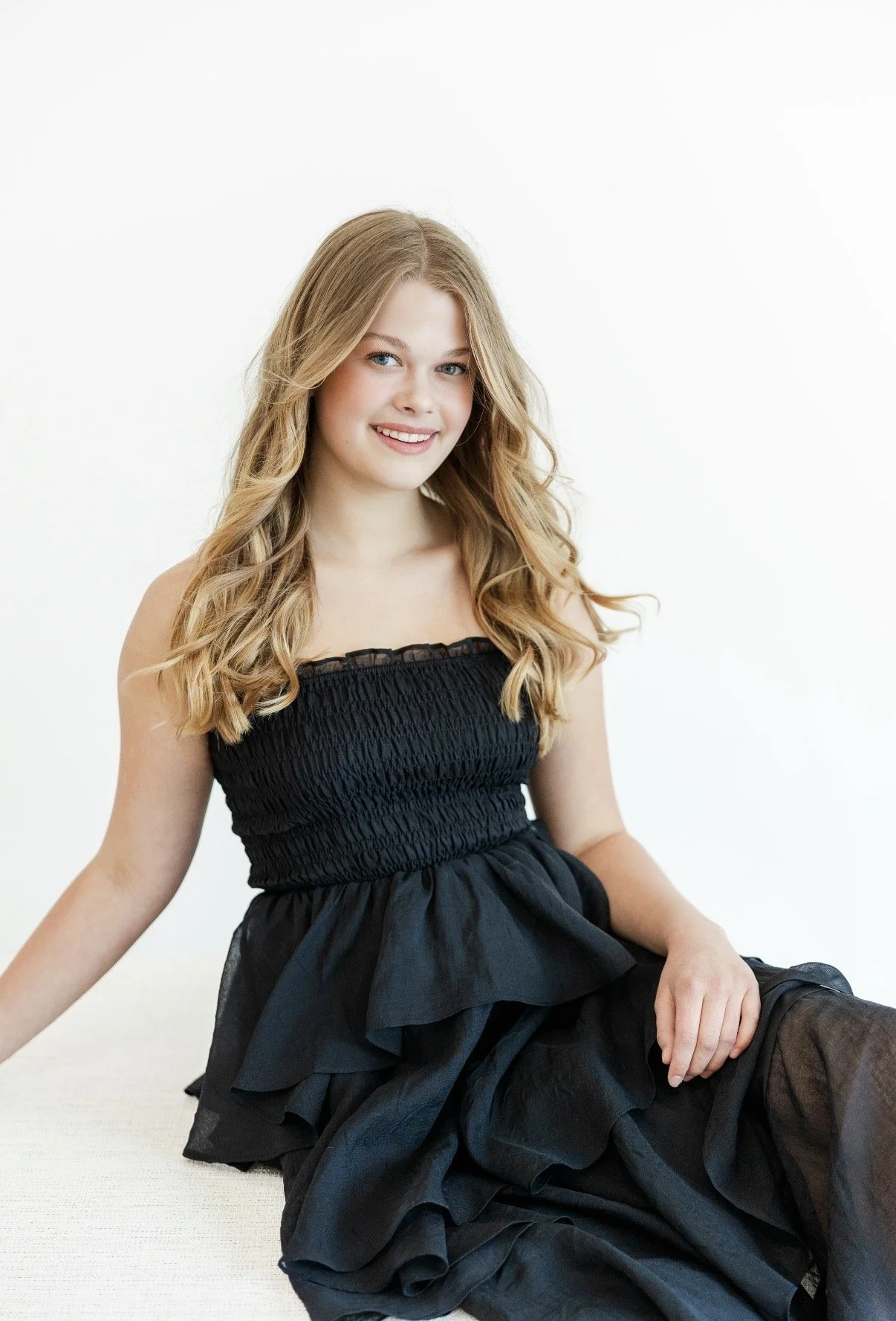 A young woman with long, wavy blonde hair and fair skin, wearing a strapless black dress, sitting on a white surface against a plain white background, smiling at the camera.