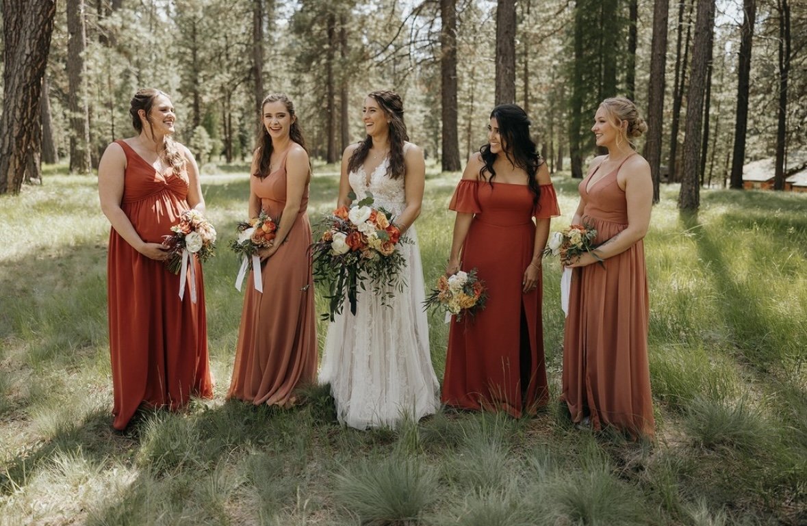 A bride and five bridesmaids standing outdoors in a forest, all holding bouquets and dressed in various shades of rust and peach.