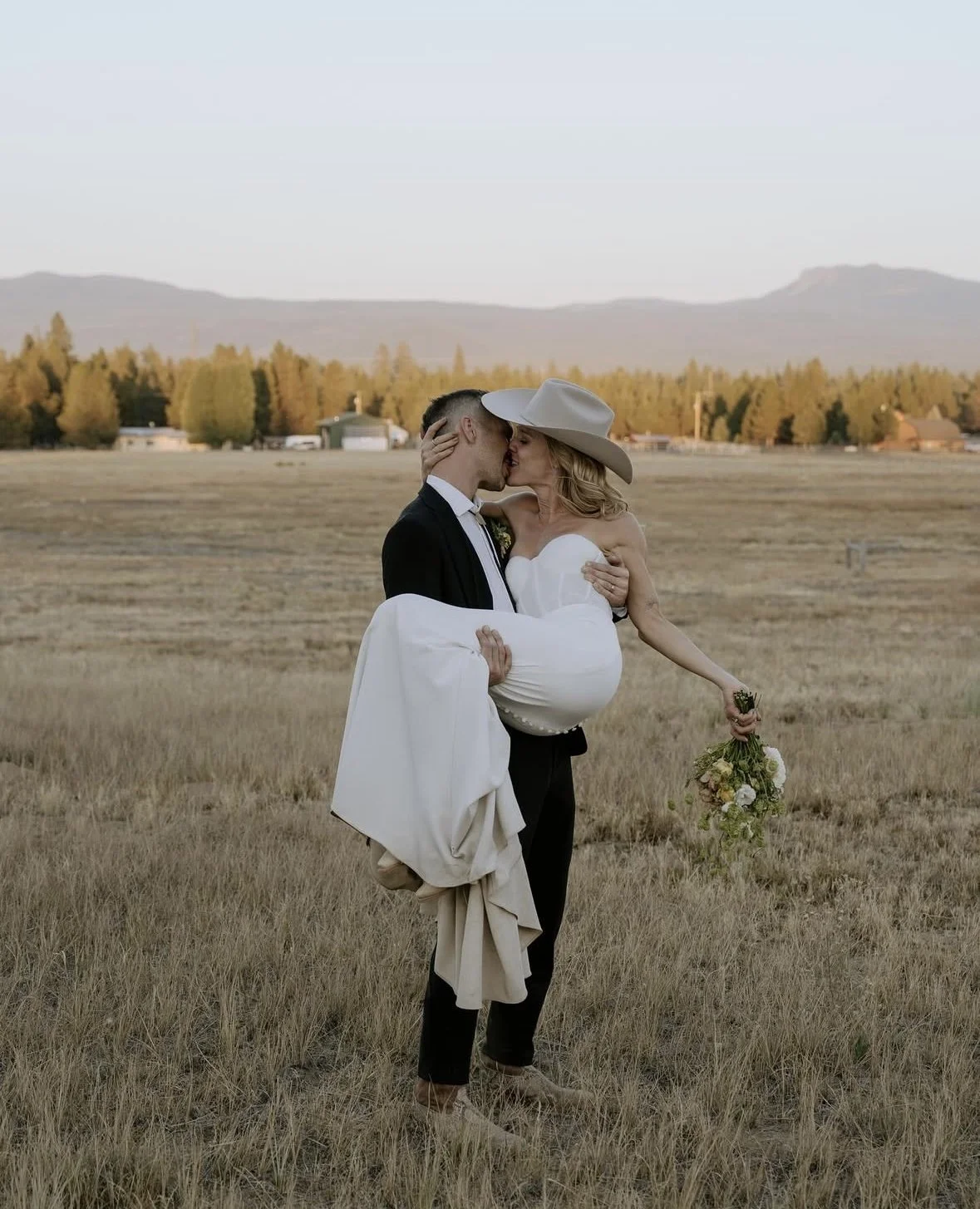 A couple in wedding attire sharing a kiss in an open field with mountains and trees in the background.