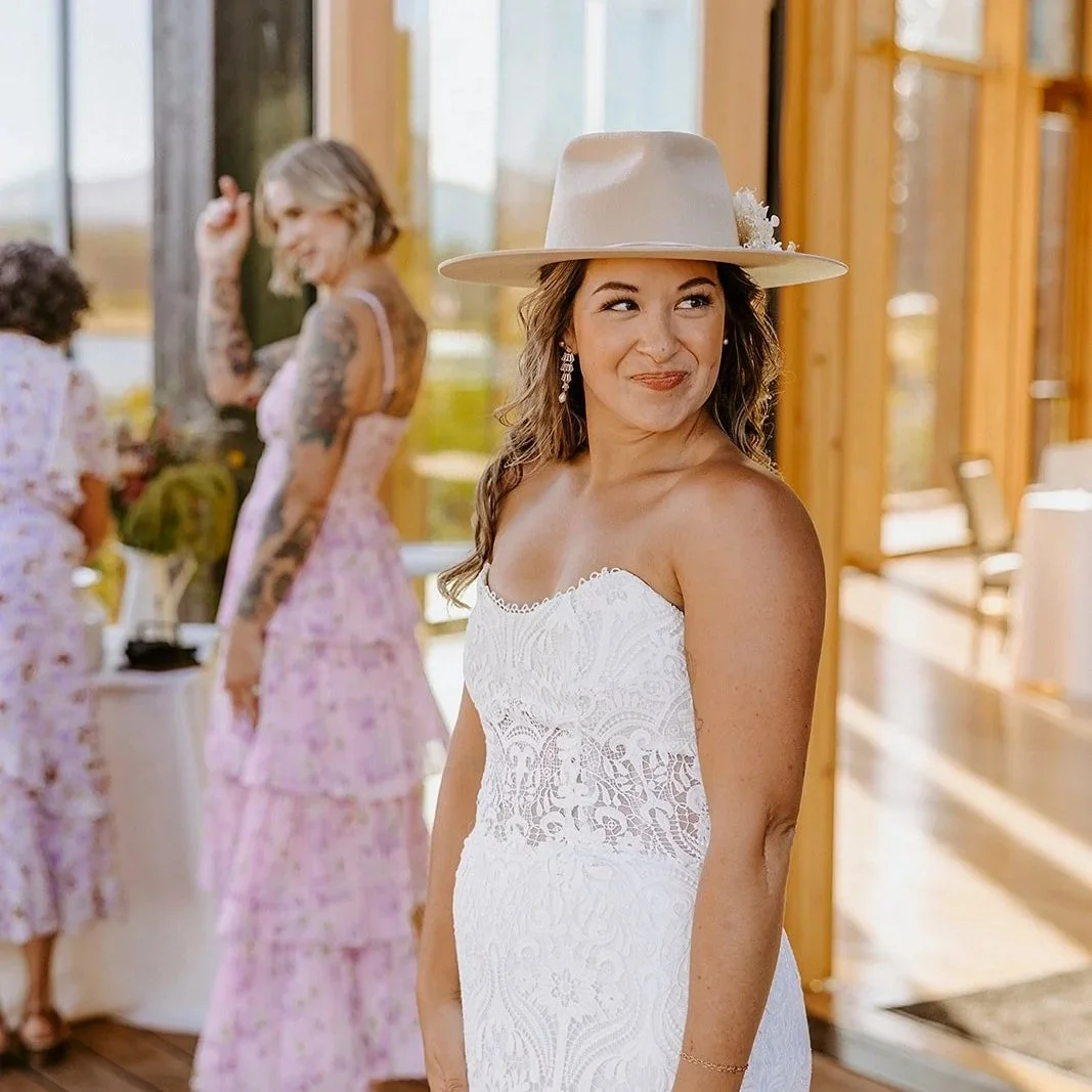 A woman in a white lace wedding dress and wide-brimmed hat smiling at a wedding reception, with others in floral dresses in the background inside a wooden structure with large windows.
