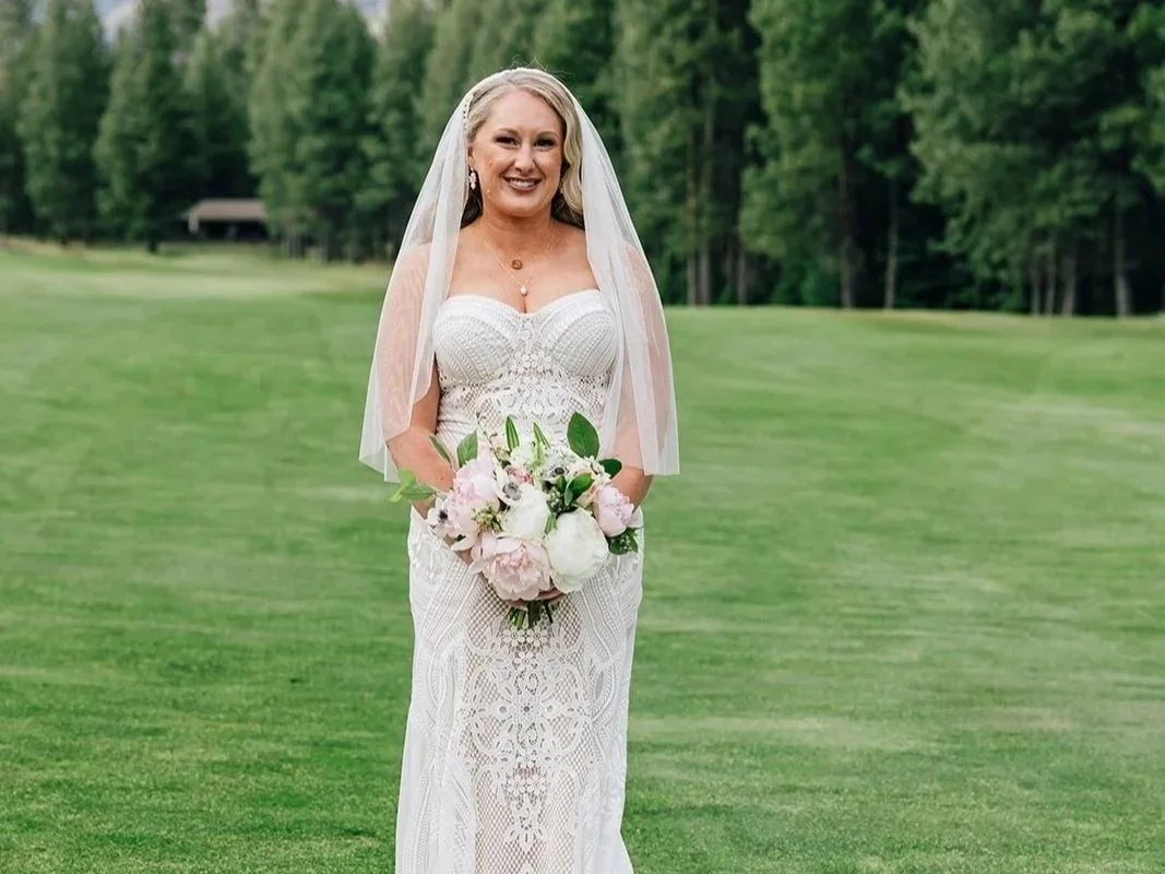 A woman in a white wedding dress holding a bouquet of pink and white flowers standing on a lush green golf course with trees in the background.