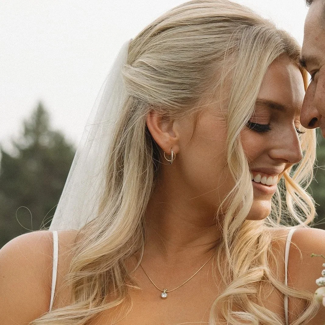 Close-up of a smiling woman with blonde hair, wearing a white dress, earrings, and a necklace, outdoors on a cloudy day.