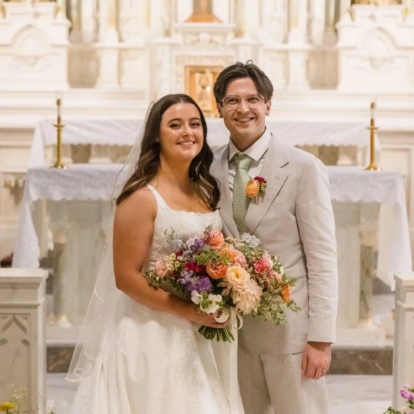 A bride and groom smiling indoors, standing closely, with the bride holding a colorful bouquet, in front of an ornate church altar.