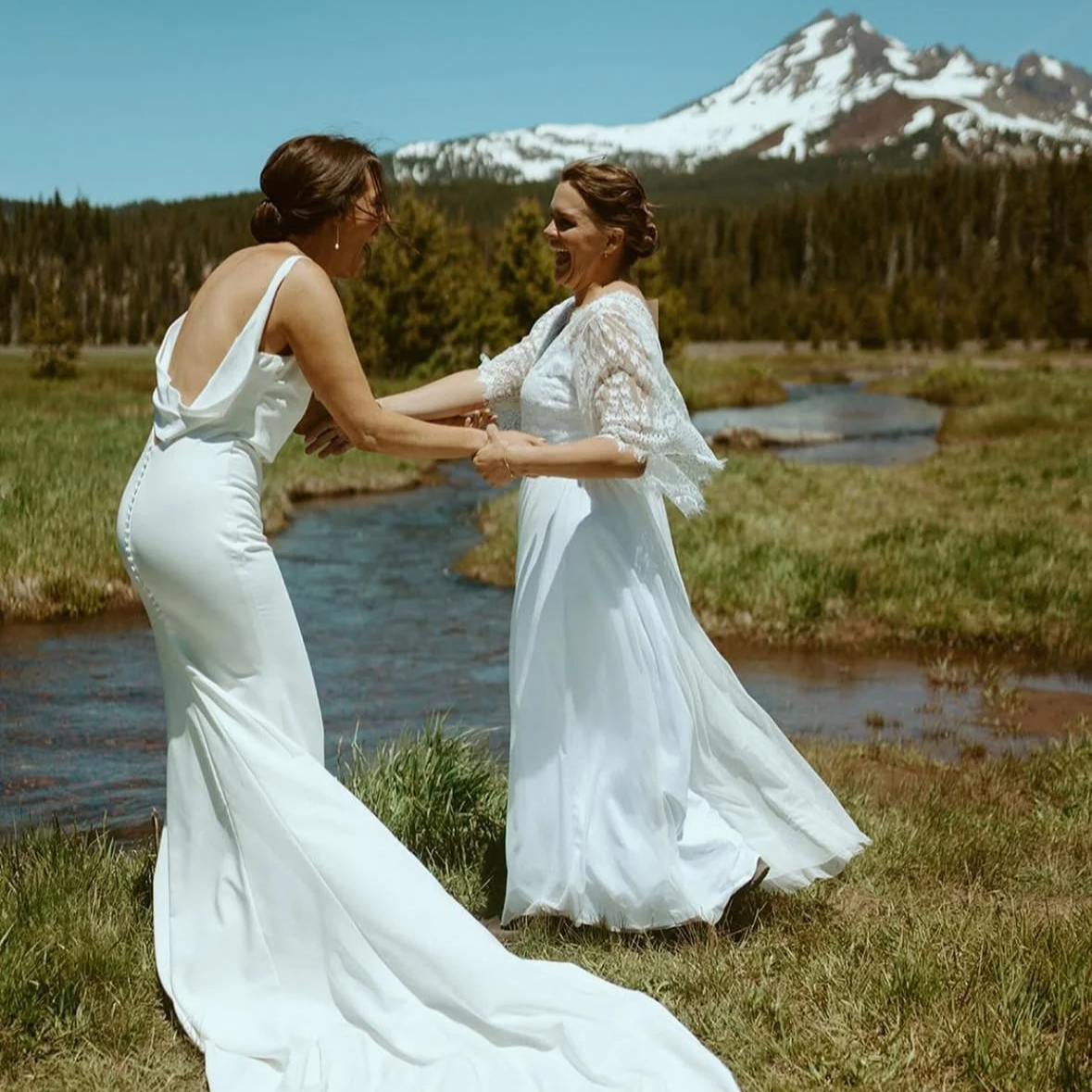 Two women in white dresses holding hands and smiling by a stream with a mountain in the background.