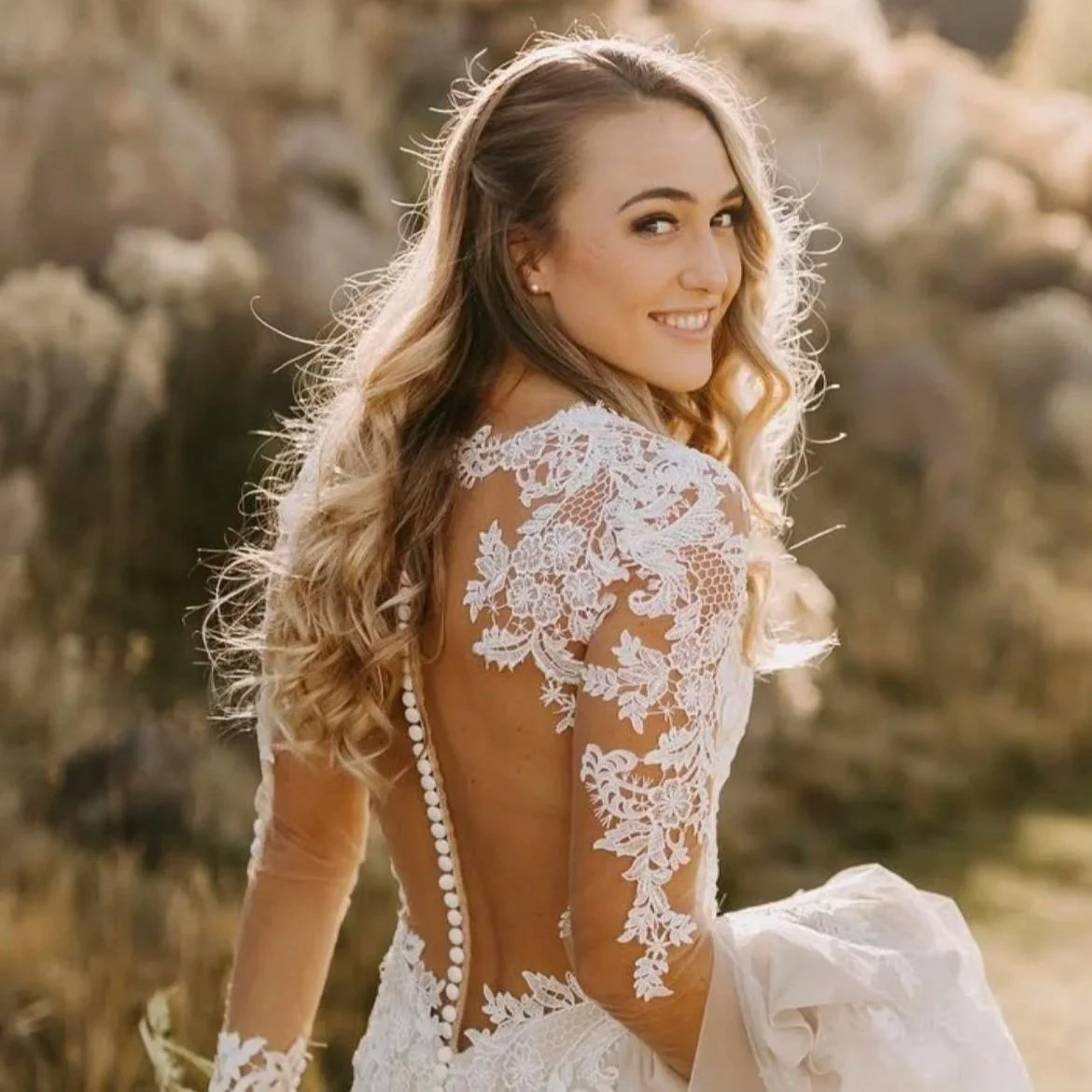 A smiling woman in a wedding dress with lace details and a low back, standing outdoors in natural surroundings.