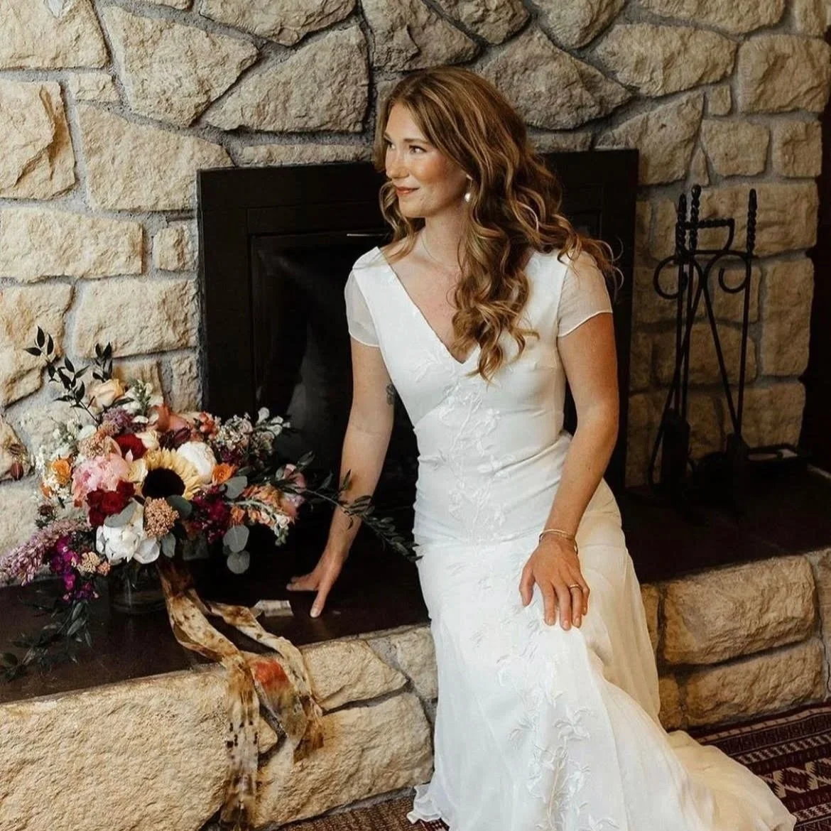A woman in a white wedding dress sitting by a stone fireplace with a bouquet of flowers.