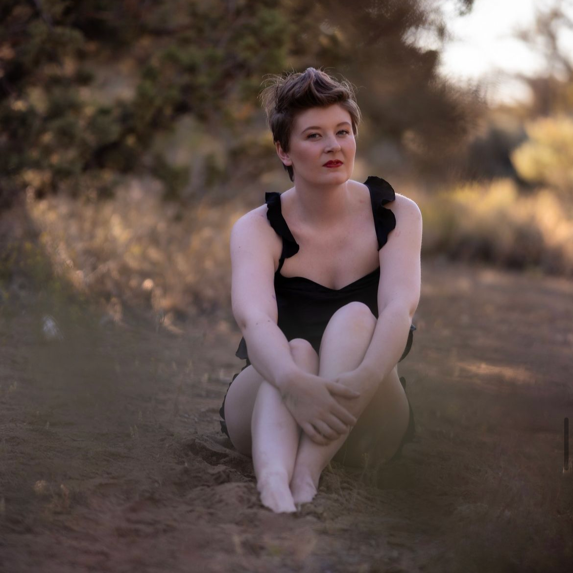 A woman with short, tousled hair and red lipstick is sitting on the ground outdoors with her knees drawn up to her chest. She is wearing a black dress with ruffled straps and has a contemplative expression, surrounded by a blurred natural background 