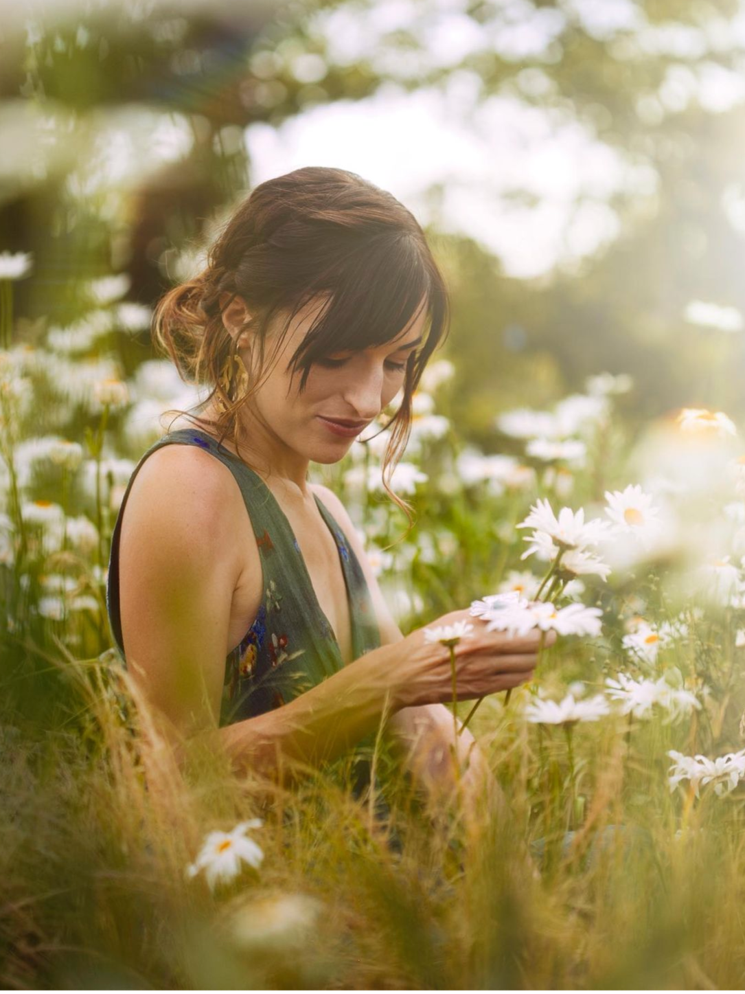 A woman with dark hair, wearing a patterned dress, is standing in a field of white daisies, looking down at the flowers she is holding.