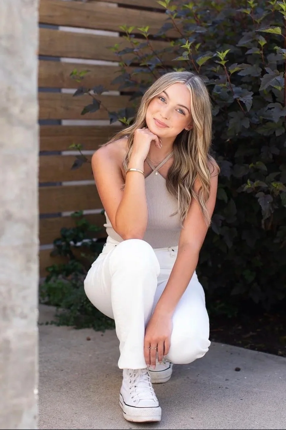 A young woman with long, wavy blonde hair kneels outdoors, smiling at the camera. She wears a beige sleeveless top, white pants, and white sneakers, with a wooden fence and leafy shrubbery behind her.