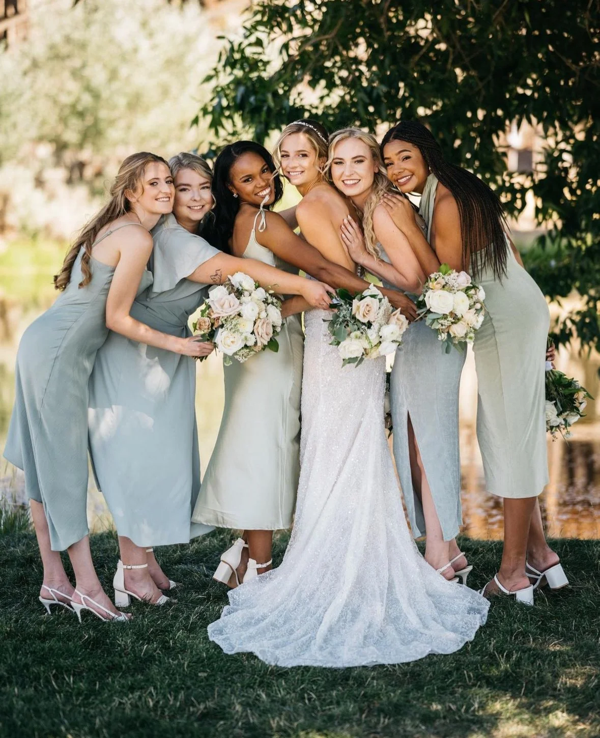 Group of seven women in elegant dresses at a wedding outdoors, holding bouquets of white and blush flowers, standing on grass with trees in the background.