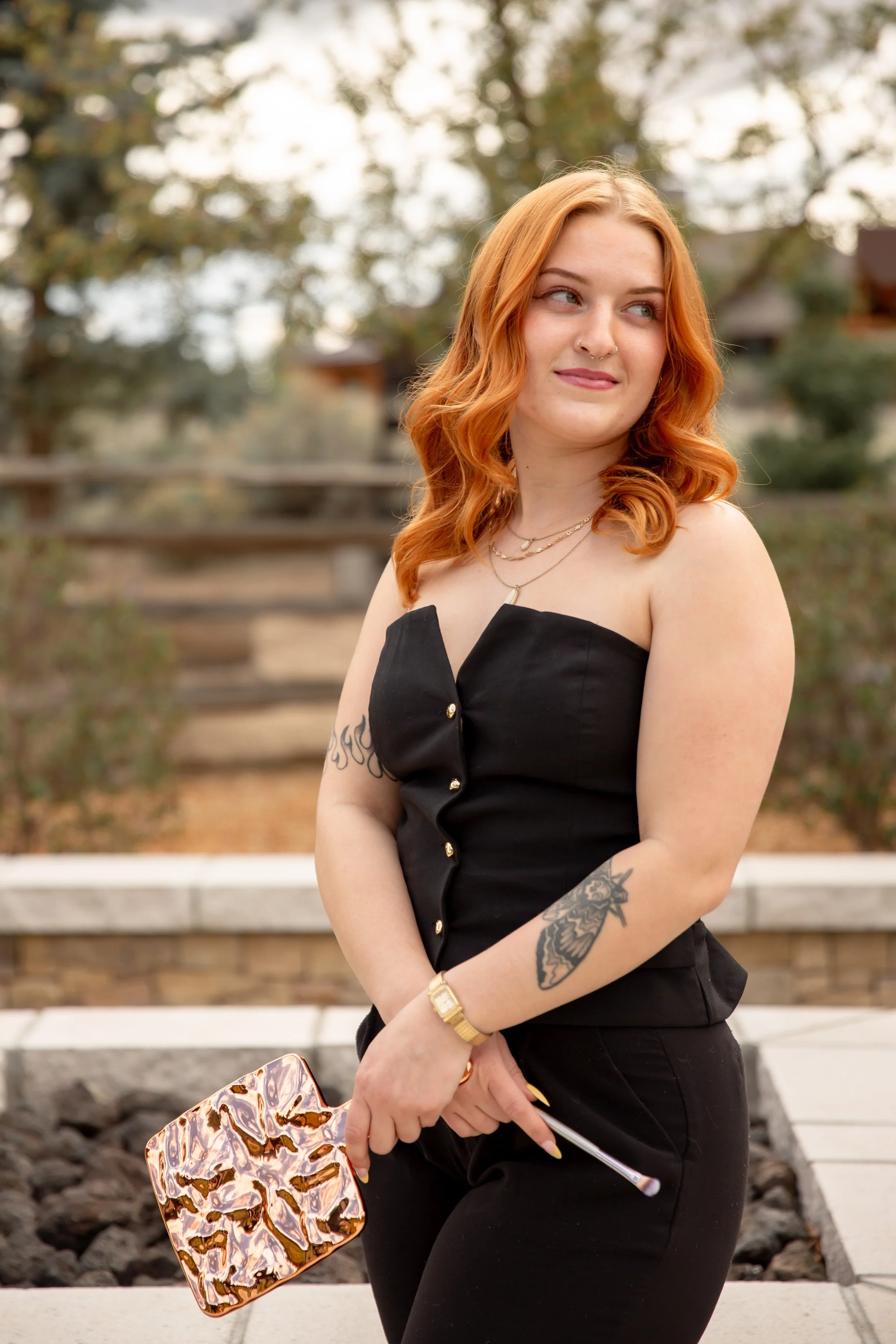 A young woman with long, wavy red hair, wearing a black sleeveless dress, holding a leopard print clutch, standing outdoors near a wooden railing with trees in the background.