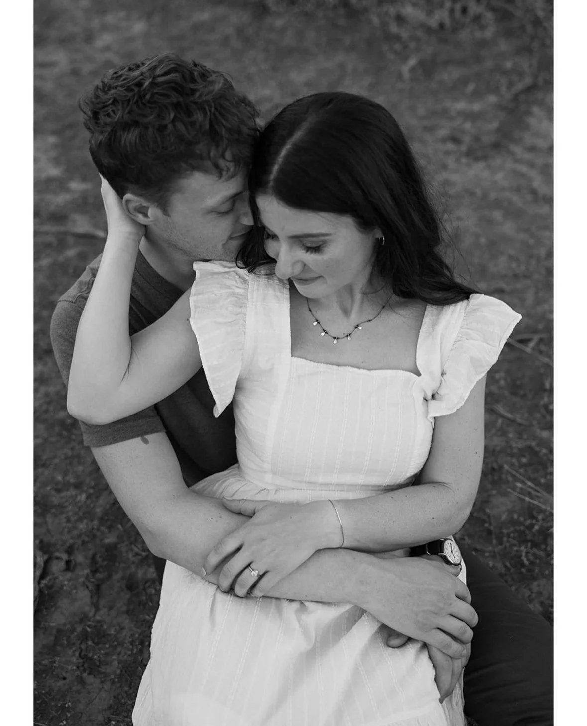 A black-and-white photo of a couple embracing outdoors, with the man's face close to the woman's forehead and her hands gently resting on his arms. The woman wears a light-colored dress with ruffled sleeves and the man wears a t-shirt. They appear af