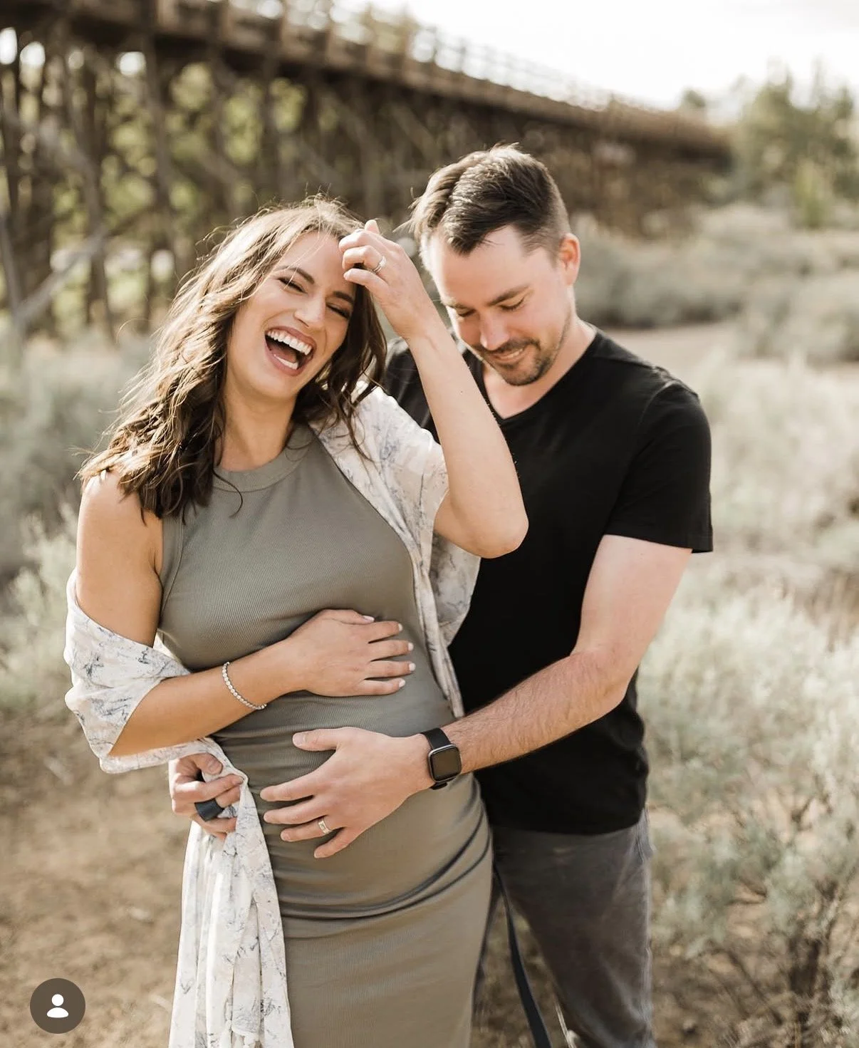 A joyful couple outdoors with the woman showing off a pregnancy bump, both smiling and holding her belly, in a natural setting with a bridge in the background.