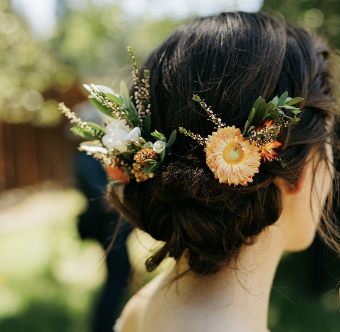 Close-up of a woman's head with dark hair styled in an elegant updo, decorated with a floral hairpiece consisting of white, orange, and yellow flowers, and green foliage, outdoors in sunlight.