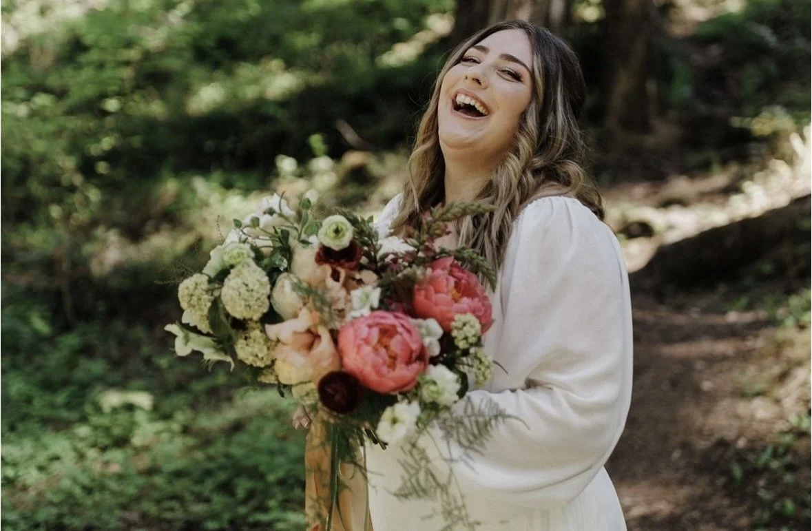 A woman with long wavy hair smiling and holding a colorful bouquet of flowers outdoors in a wooded area.