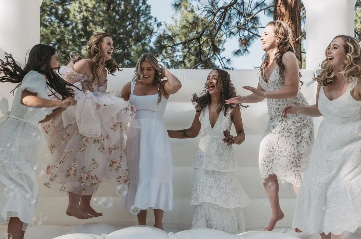 Group of seven women in white dresses having fun and dancing outdoors on a sunny day, with bubbles floating around.