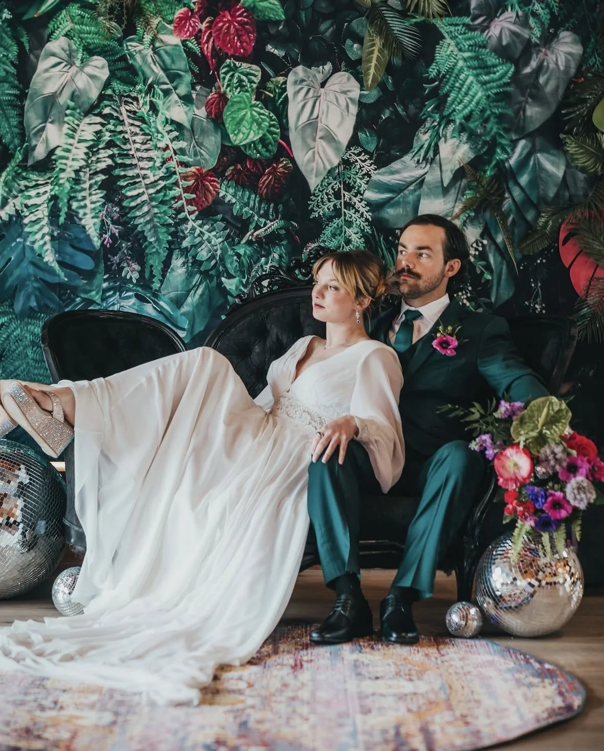 A bride and groom sitting on a black vintage sofa with a lush green tropical leaf background, a large bouquet of colorful flowers on the right, and disco balls on the floor.