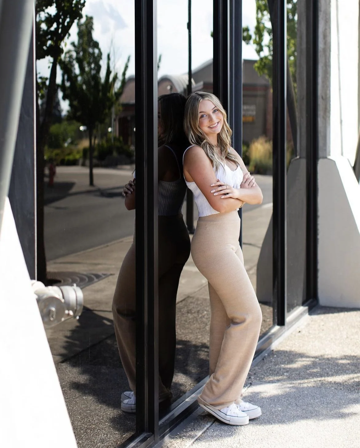 A young woman with long blonde hair, wearing a white sleeveless top and beige pants, stands outside near a glass door, smiling with her arms crossed. Her reflection is visible in the glass.