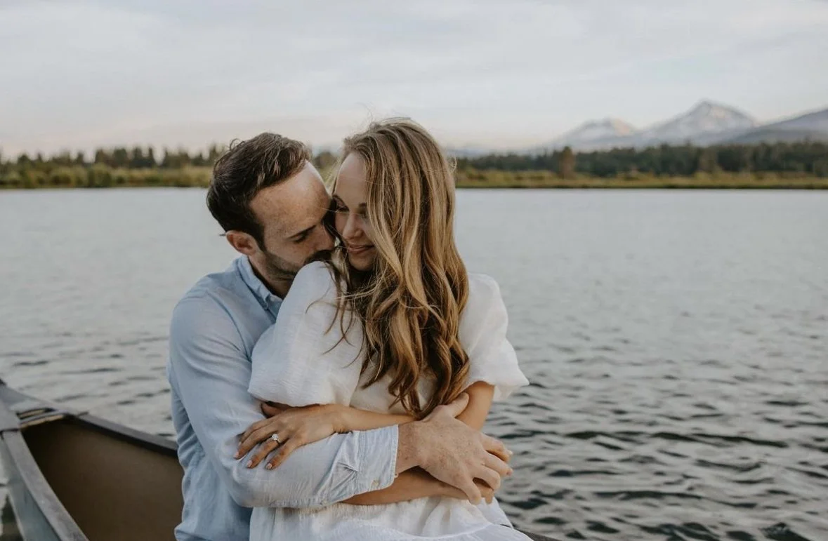 A couple sitting by a lake, embracing, with mountains in the background, under a cloudy sky.