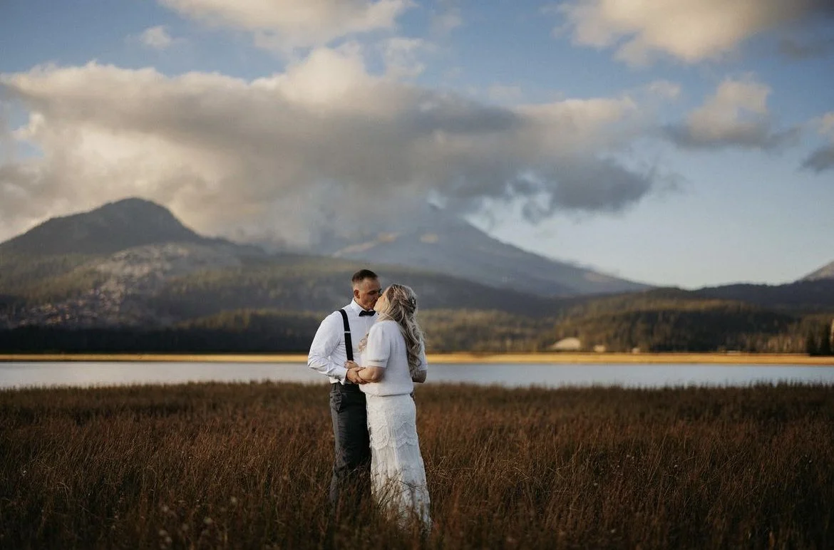 A couple standing closely together in a field next to a lake with mountains in the background, during sunset or late afternoon.