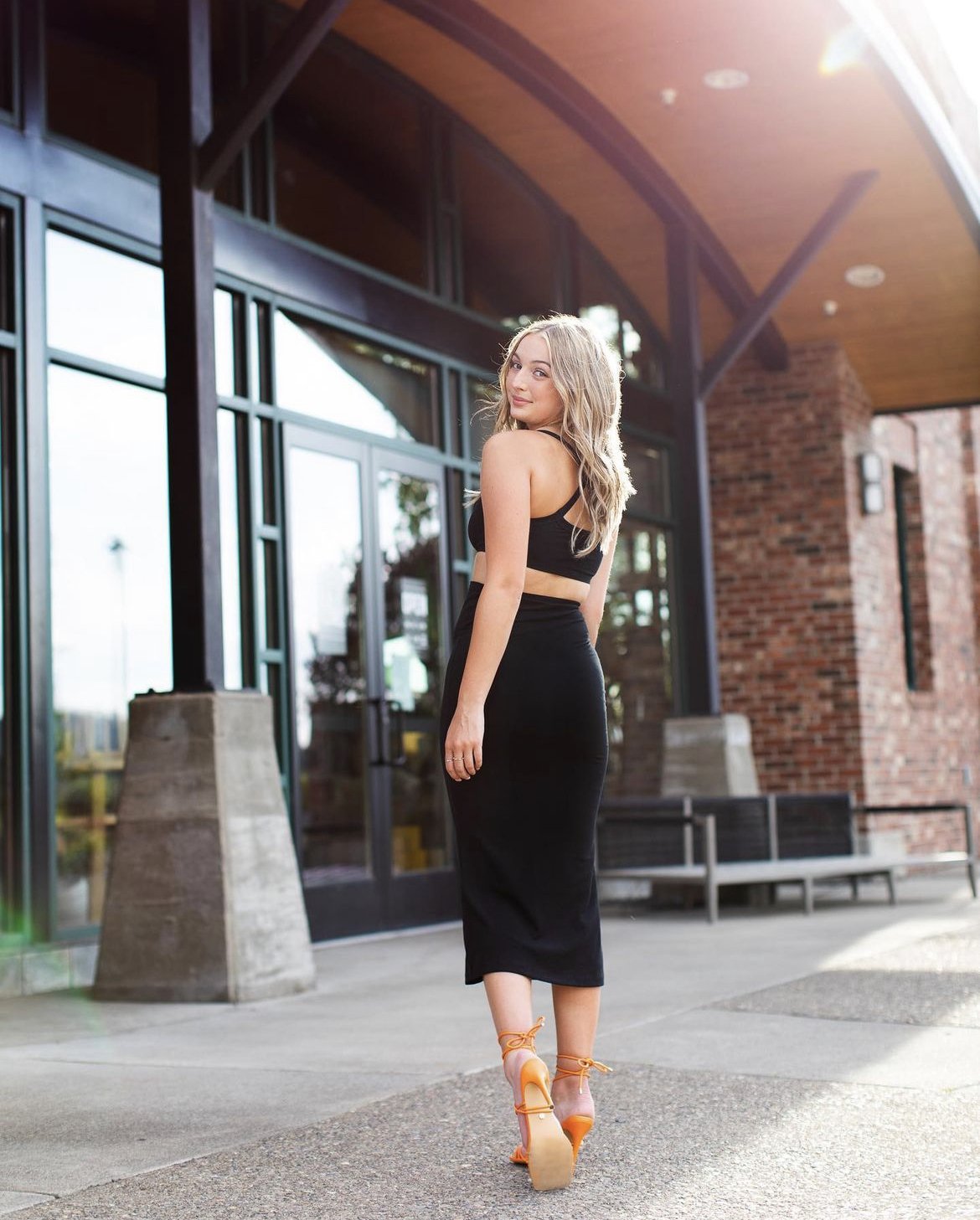 A woman in a black crop top and high-waisted black skirt stands outdoors near a building with large glass windows, wearing orange high heels, smiling at the camera.
