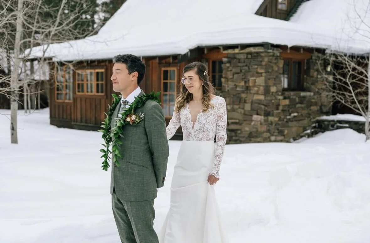 A bride and groom standing outdoors in the snow, with the bride pulling the groom's arm, in front of a rustic cabin.