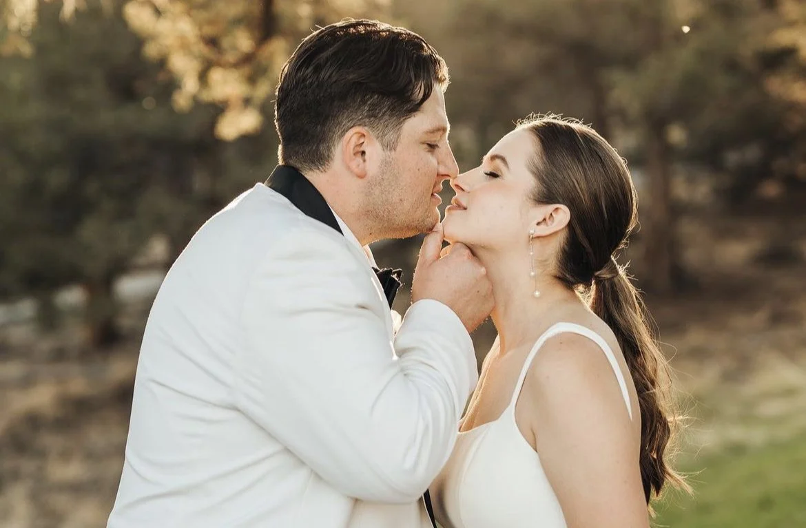 A bride and groom about to kiss outdoors at sunset, with the groom wearing a tuxedo and the bride in a white wedding dress.