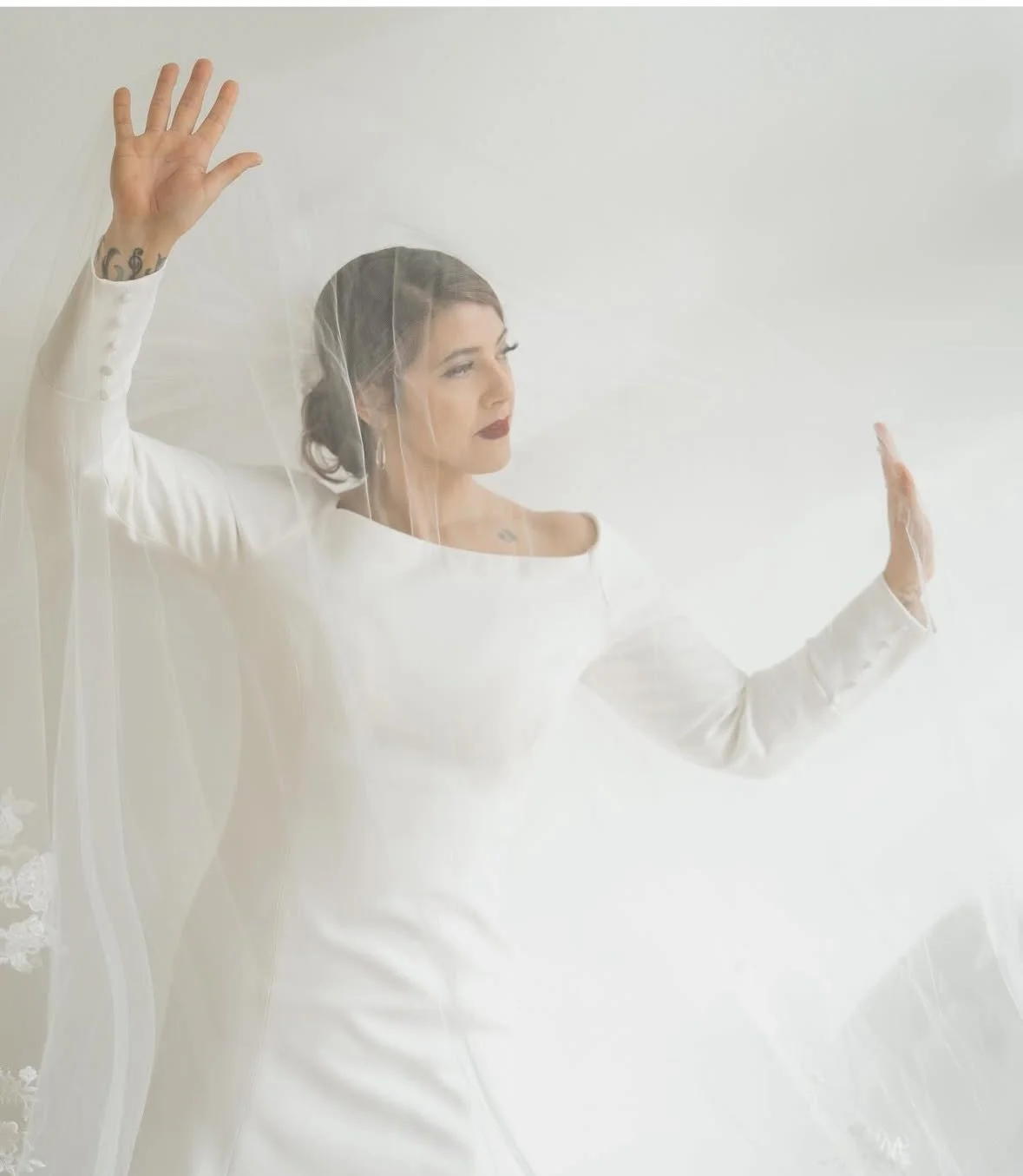 A woman in a white wedding dress and veil with arms raised, standing behind a glass or plastic panel, looking to her right against a plain light-colored background.
