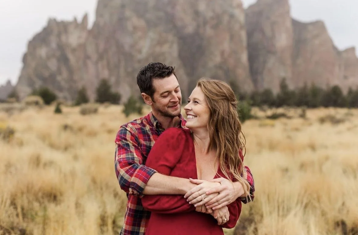 A smiling couple embracing in a field with mountains in the background.