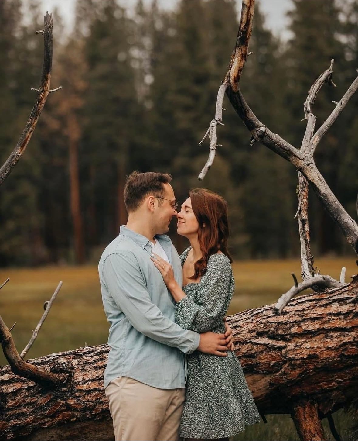 A young couple standing close together outdoors, touching noses and smiling, with a fallen tree and forest background.