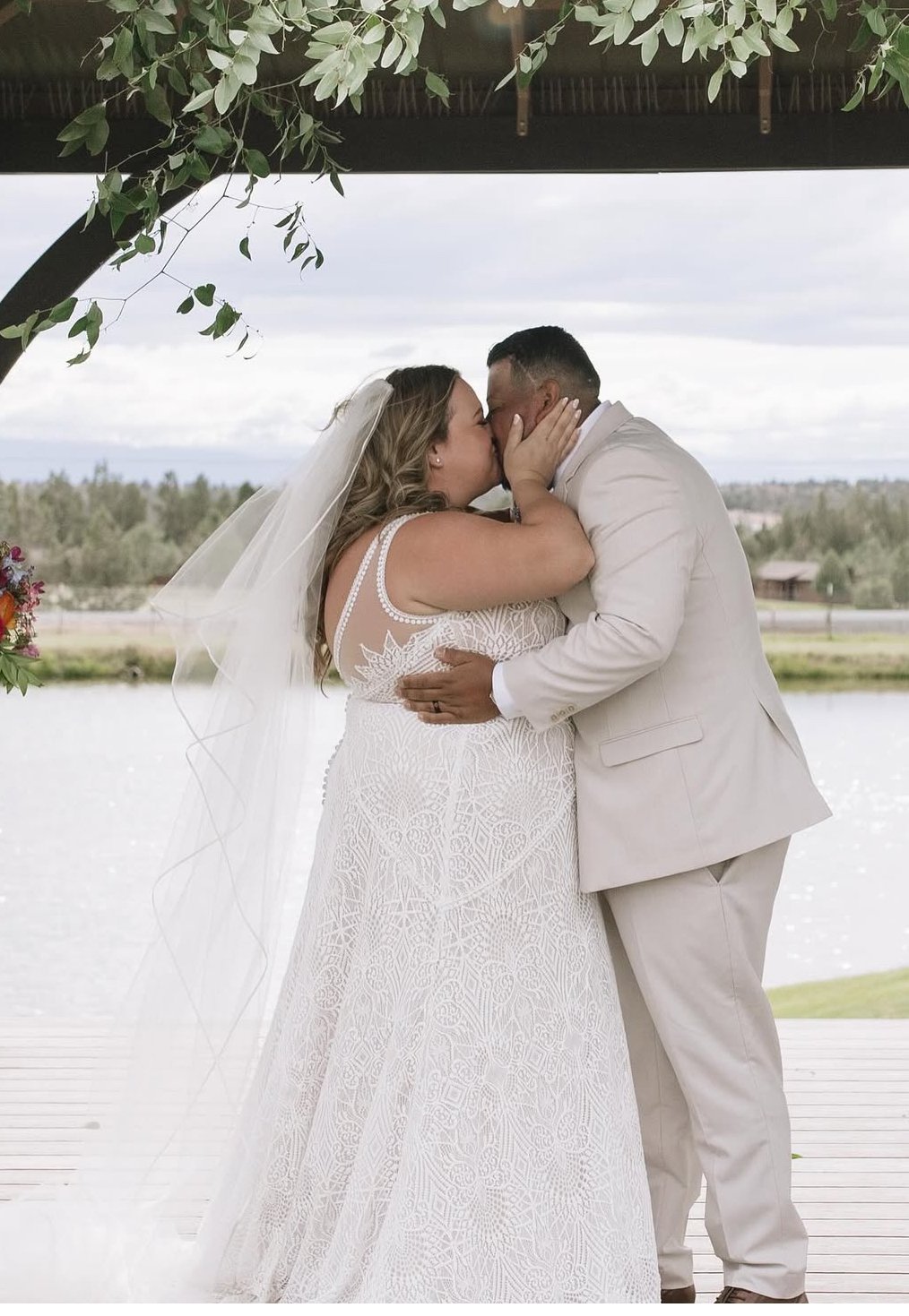 A bride and groom share a kiss during their wedding ceremony outdoors near a body of water, with trees and a cloudy sky in the background.