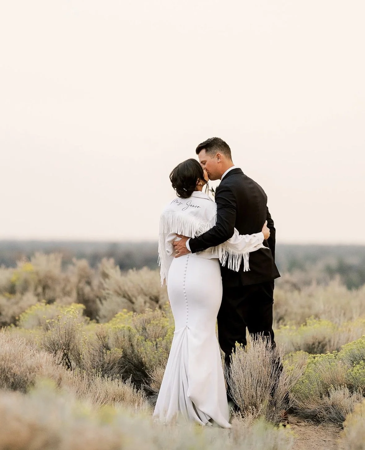 A couple dressed in wedding attire sharing a kiss in an outdoor field with bushes and trees in the background at sunset.